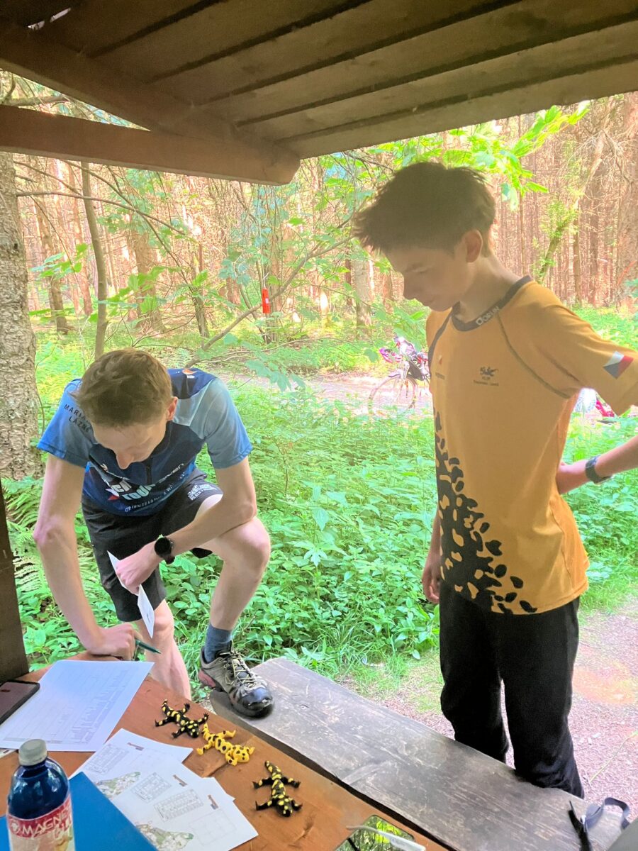 dvě osoby ve sportovním oblečení hledící na mapy umístěné na stole a vedle map ležící plastové mloky